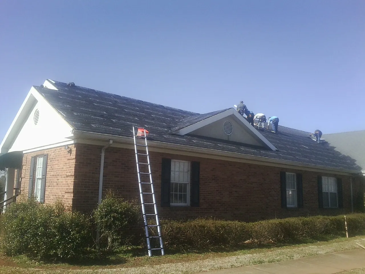 Crew tearing shingles off a brick ranch roof with a ladder set — Emergency Roof Repair work by our crew in Flatonia